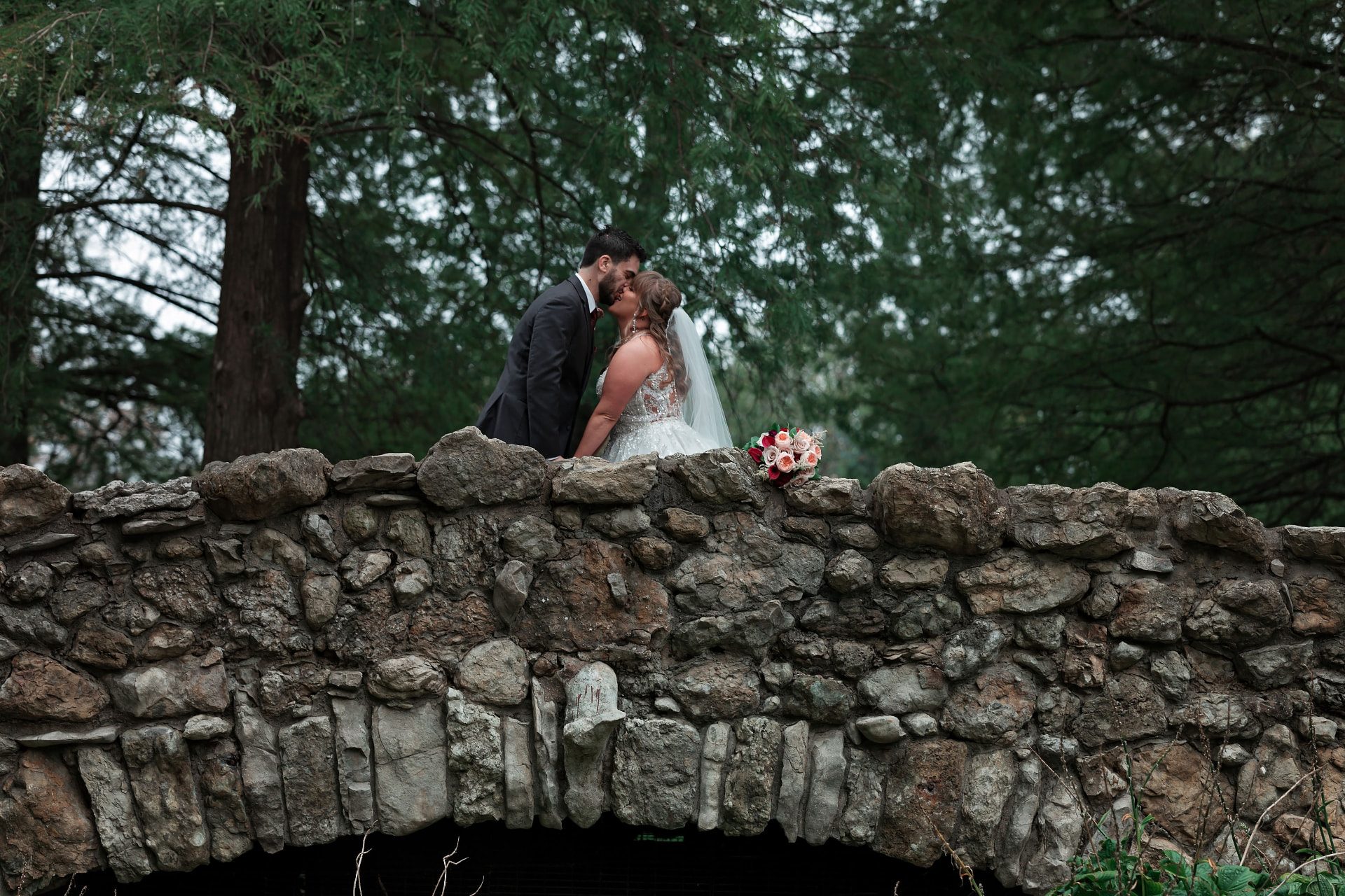 Bride and groom kissing across a stone arch bridge bordered by dense evergreens