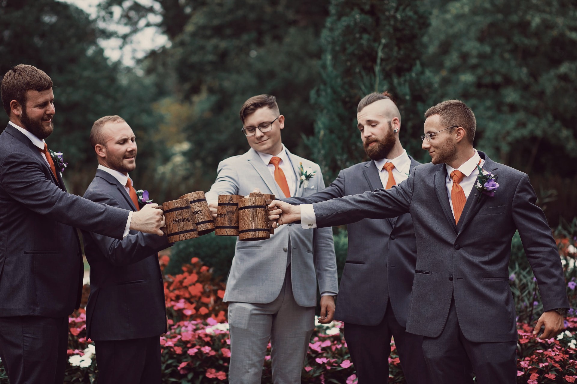 Groomsmen in gray suits toasting with carved wooden mugs among the flowers