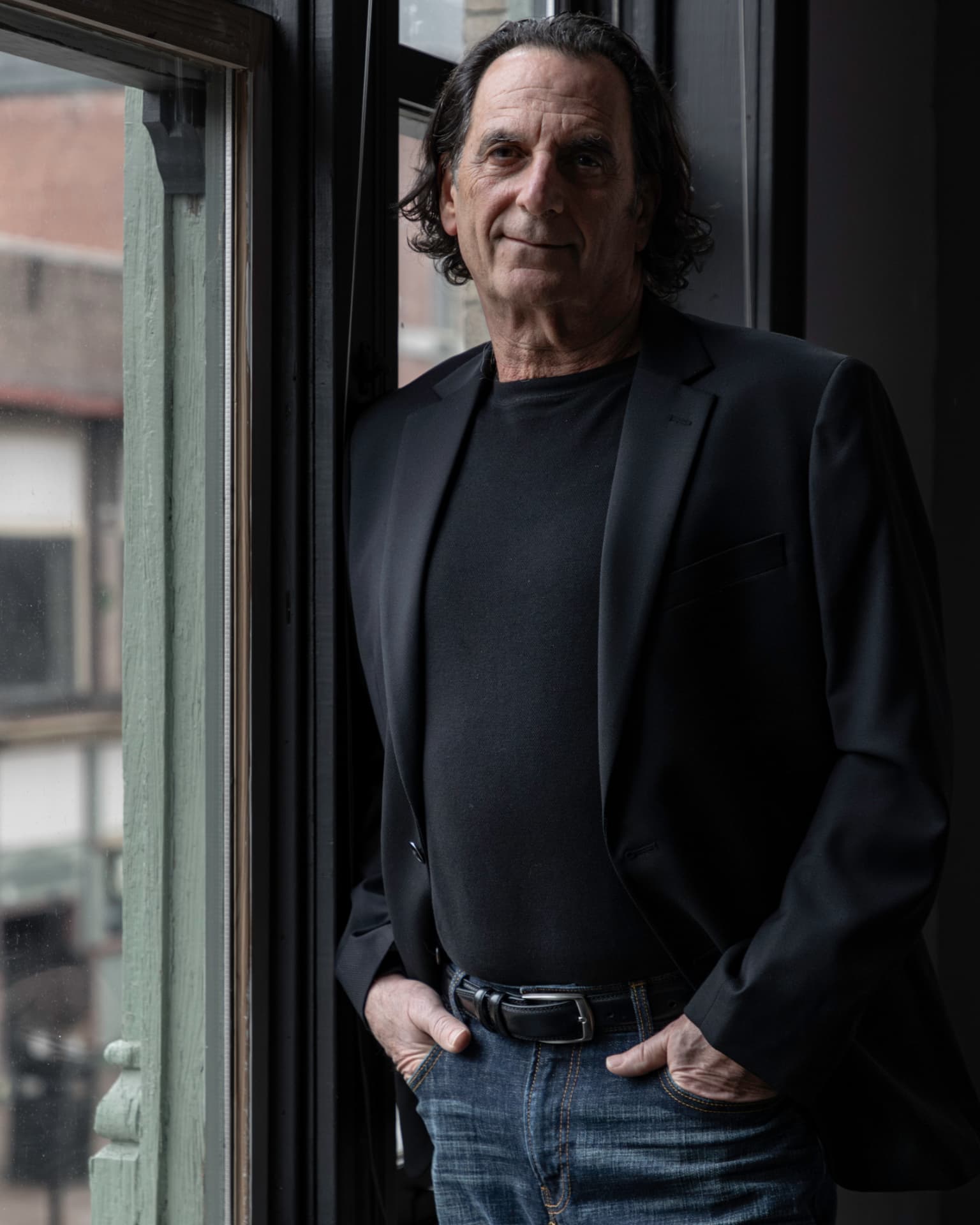 Man in a black blazer leaning against a tall window in soft daylight