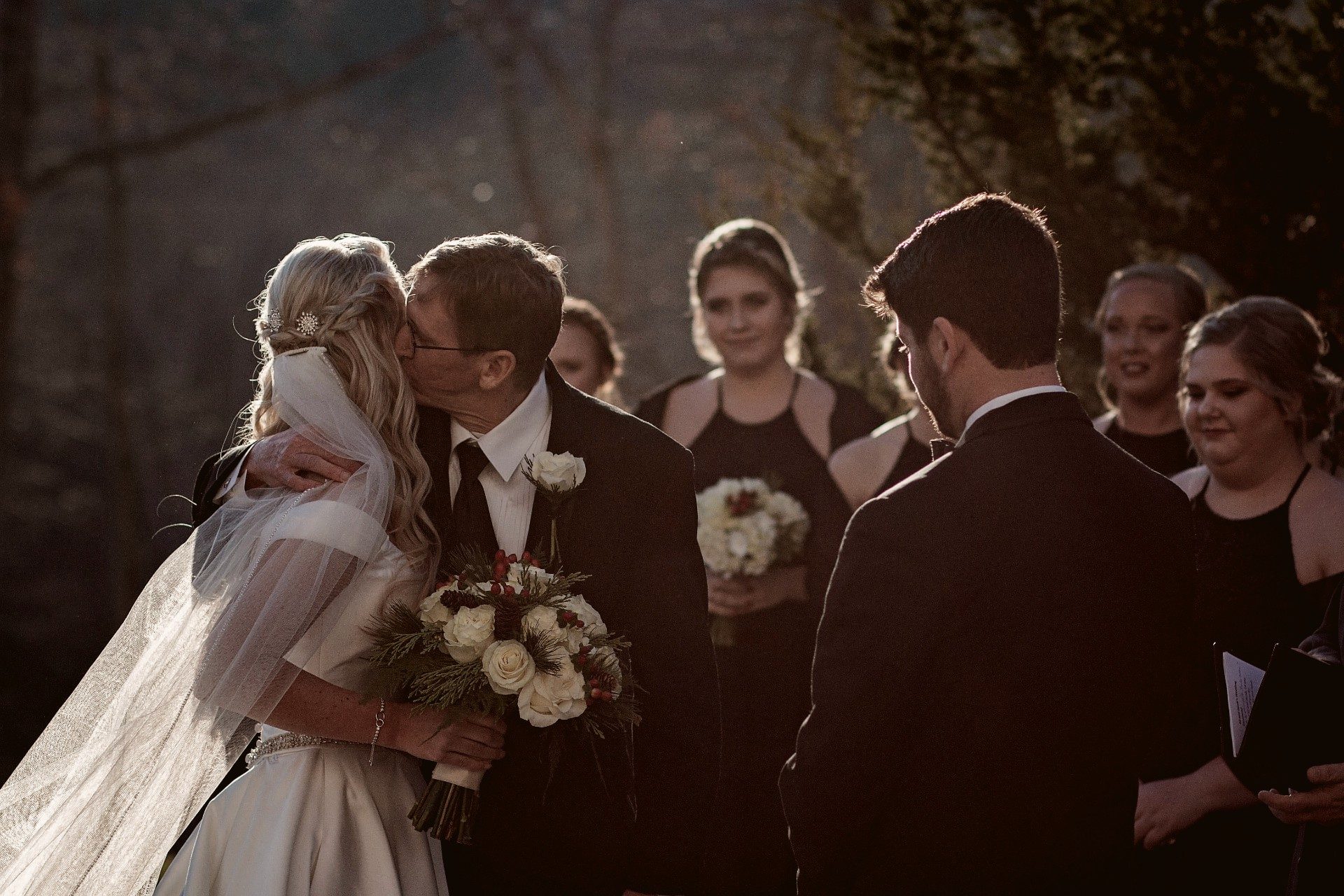 Father kissing his daughter's cheek during the ceremony as the groom looks on, backlit