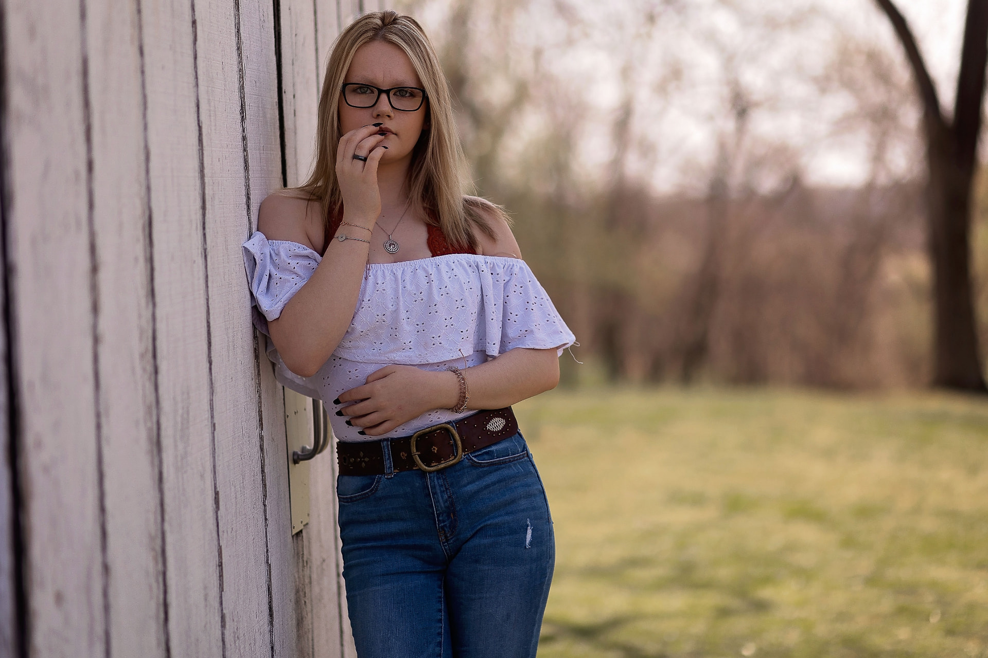 Senior in a white off-the-shoulder top leaning against a weathered white barn wall