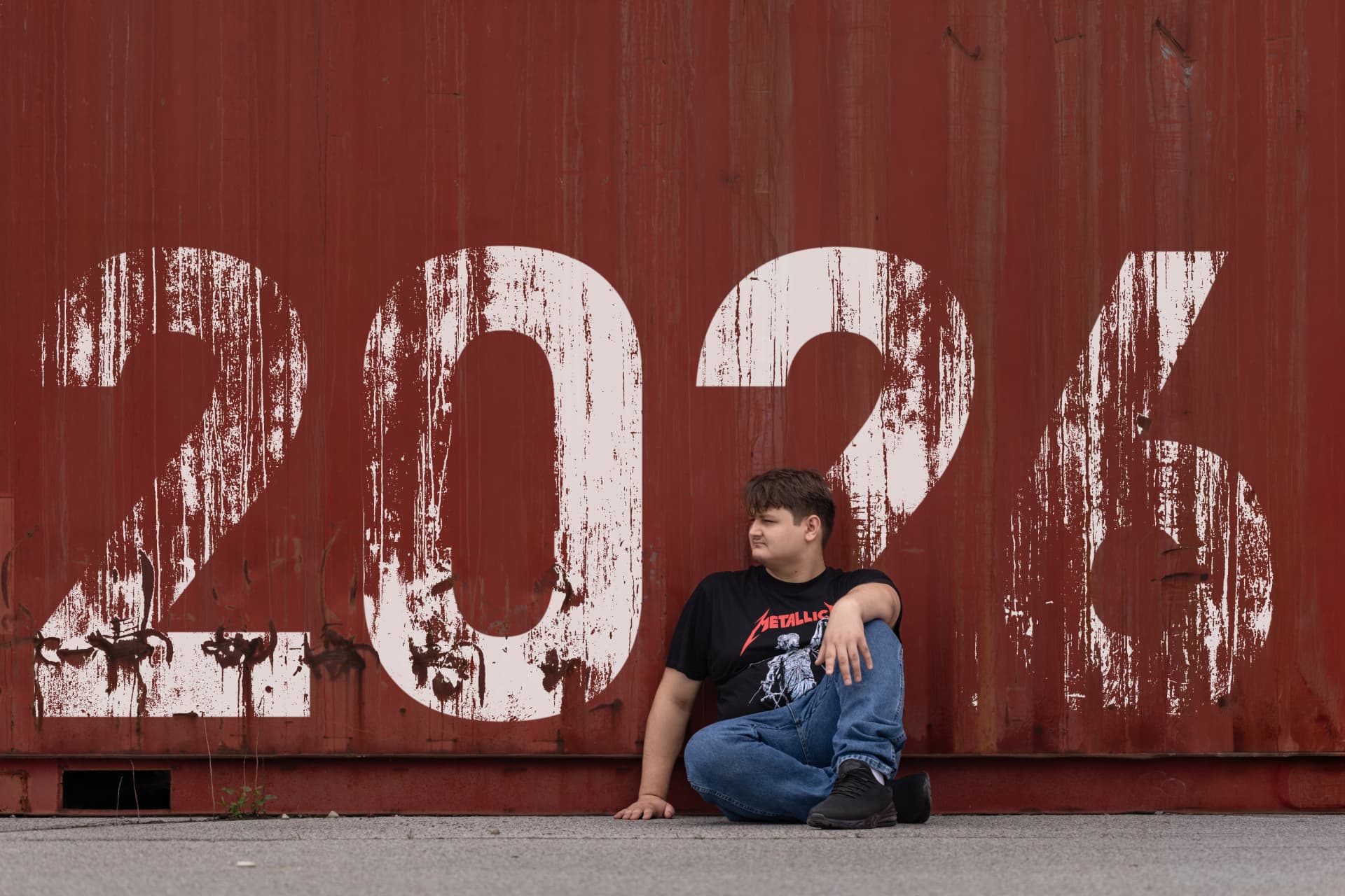 Senior in a Metallica shirt sitting cross-legged against a red shipping container marked 2026