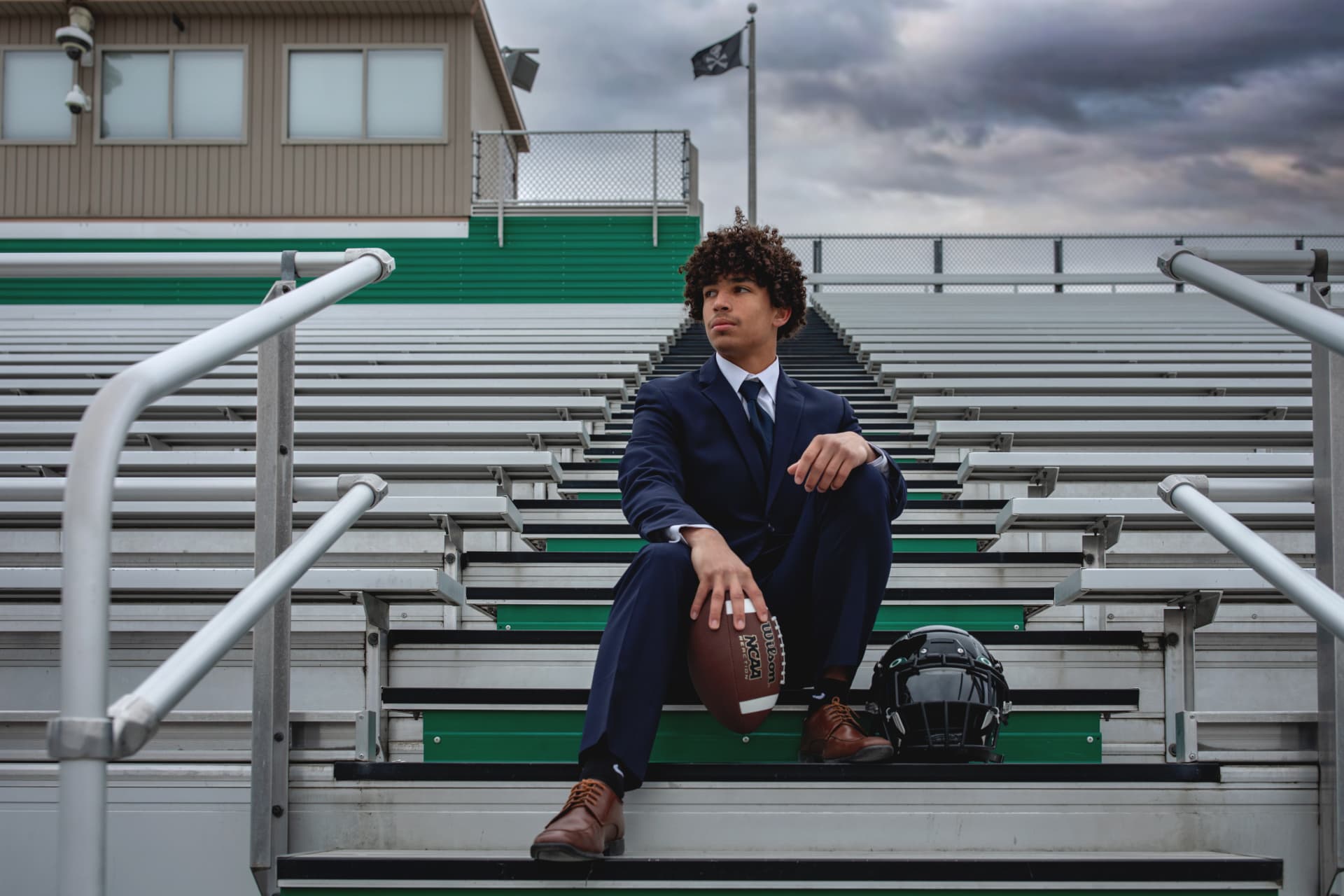 Senior in a navy suit seated on stadium bleachers with a football in hand and a helmet on the step beside him
