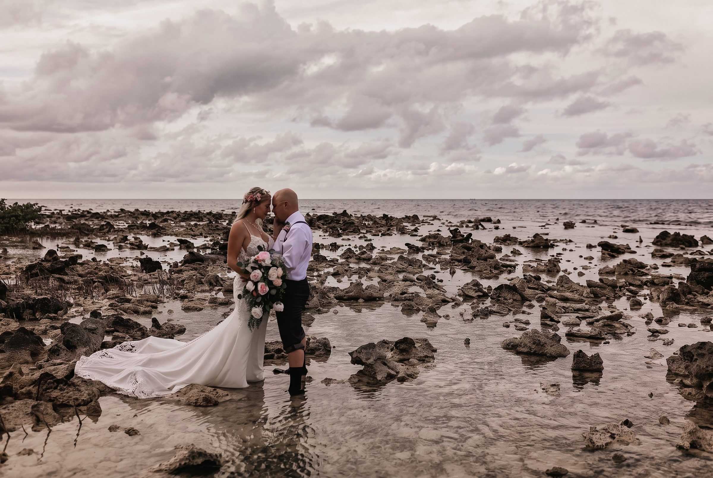 Bride and groom on a rocky shoreline beneath a cloudy sky