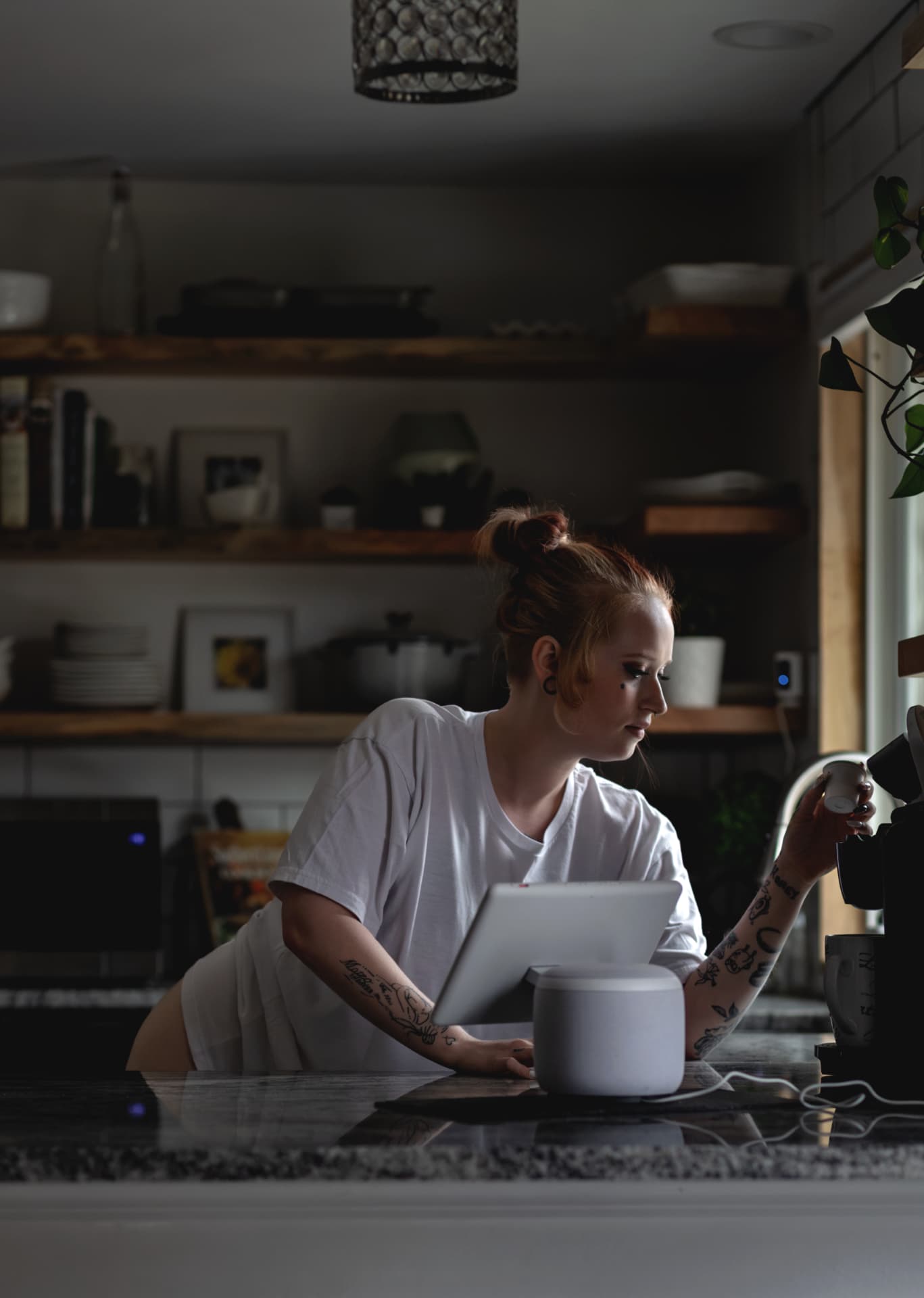 Tattooed woman in a white t-shirt making coffee in a dim kitchen, lit by window light