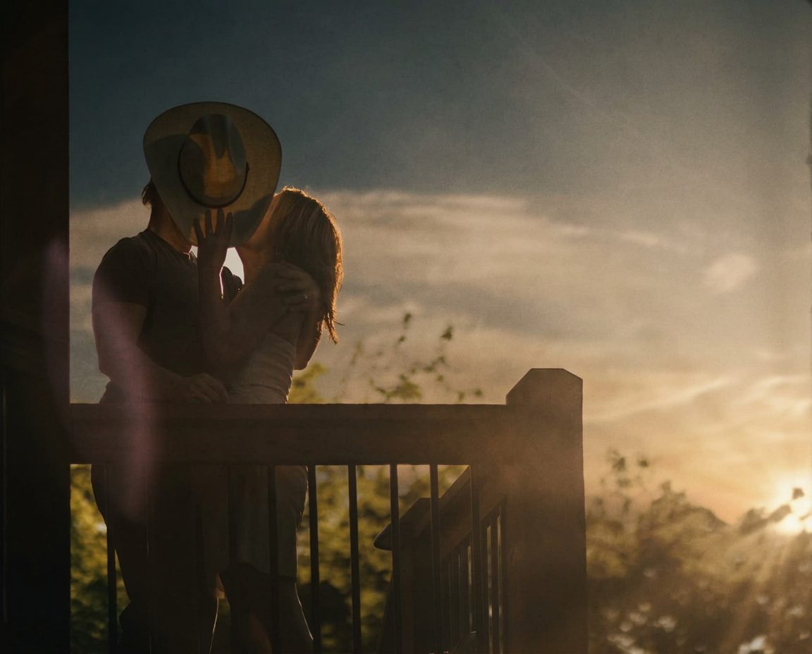 Couple kissing behind a cowboy hat on a porch at sunset