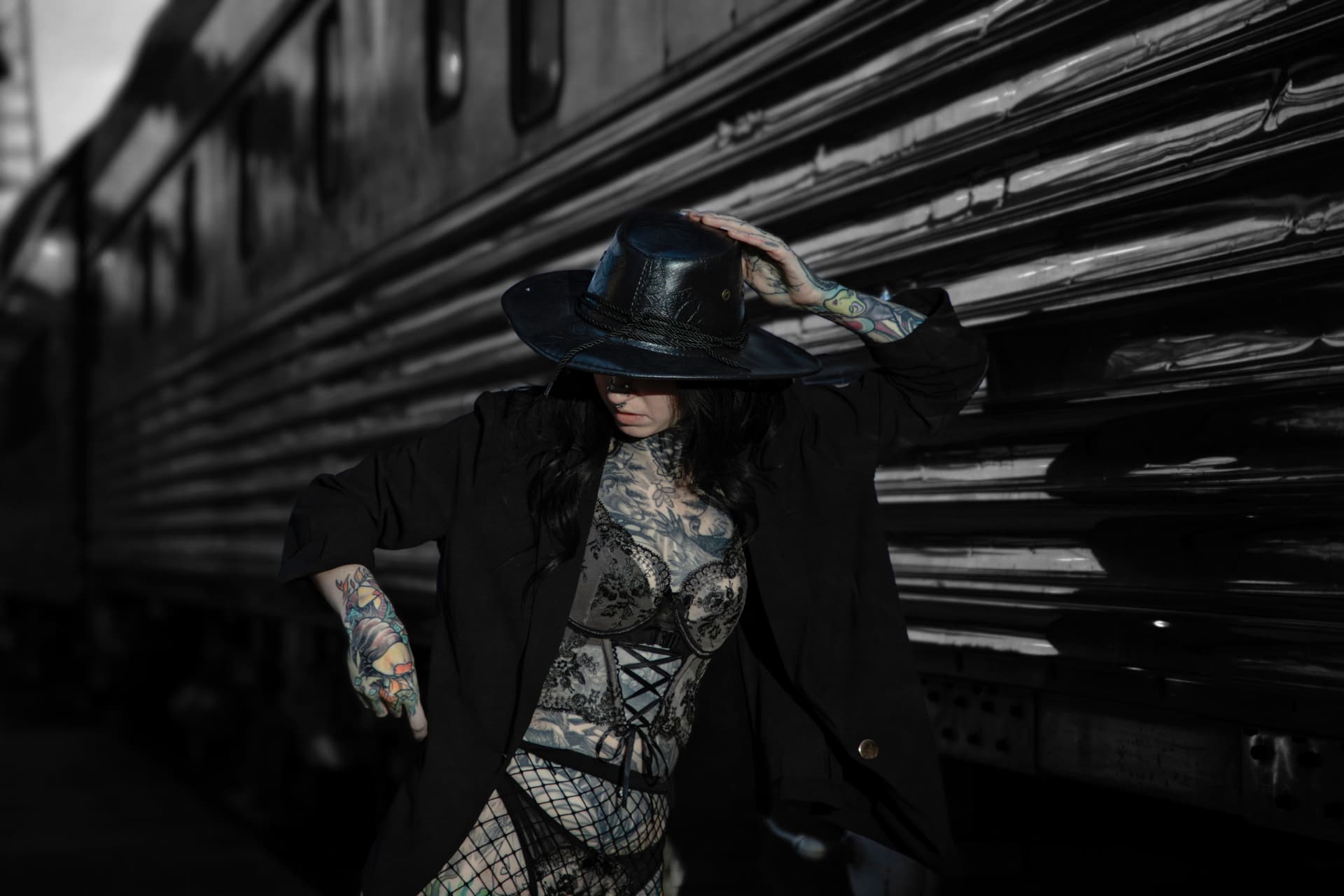 Tattooed woman in a black wide brim hat and jacket posing beside a vintage train car